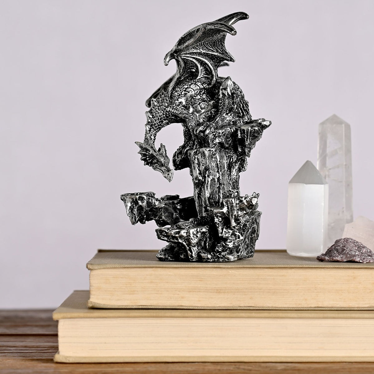 Dragon on Rocks Metallic Effect Resin Figurine in silver, perched with wings raised on a textured rocky column, shown on stacked books beside crystal decor.