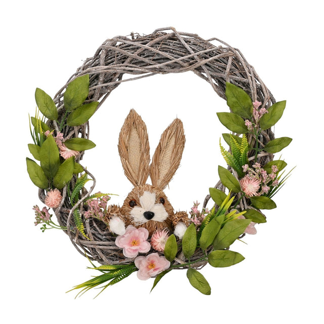 Round twig wreath with a tan straw-textured bunny peeking from the base, surrounded by pink flowers and green foliage, on a plain white background.