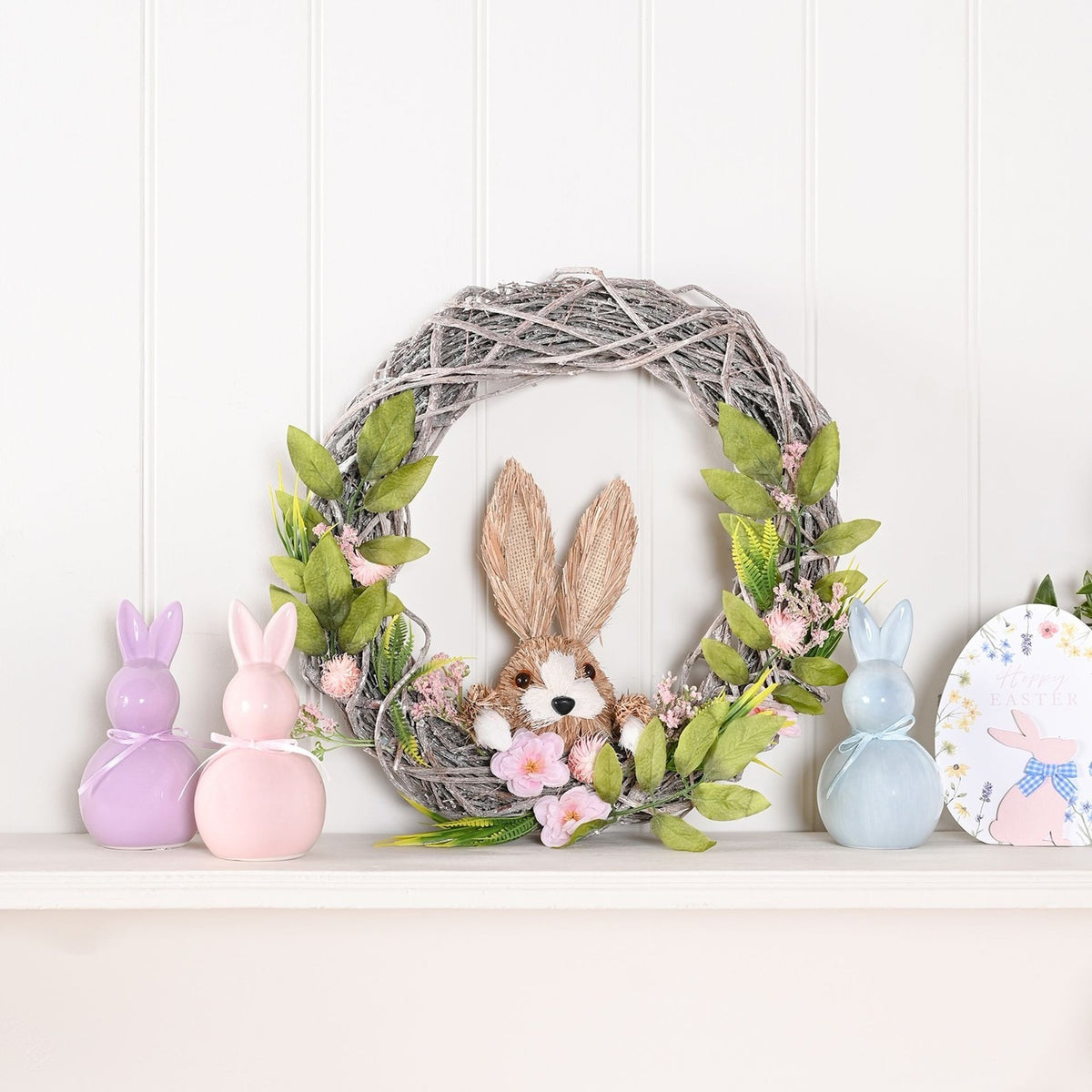 Round Easter wreath of grey-washed woven branches with a straw-textured bunny, pink flowers and green foliage, displayed on a mantel with pastel bunny figurines.
