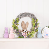 Round Easter wreath of grey-washed woven branches with a straw-textured bunny, pink flowers and green foliage, displayed on a mantel with pastel bunny figurines.