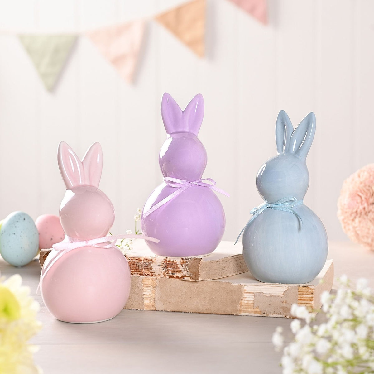 Set of 3 Pastel Coloured Easter Bunny Ceramic Figurines in pink, lilac and grey-blue, each with a glossy glaze, round body, tall ears and a matching ribbon bow, displayed on a wooden block.