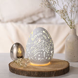 White glass Easter egg LED light-up decoration with cut-out rabbit and floral pattern, displayed glowing on a wood slice beside a vase and gypsophila sprigs.