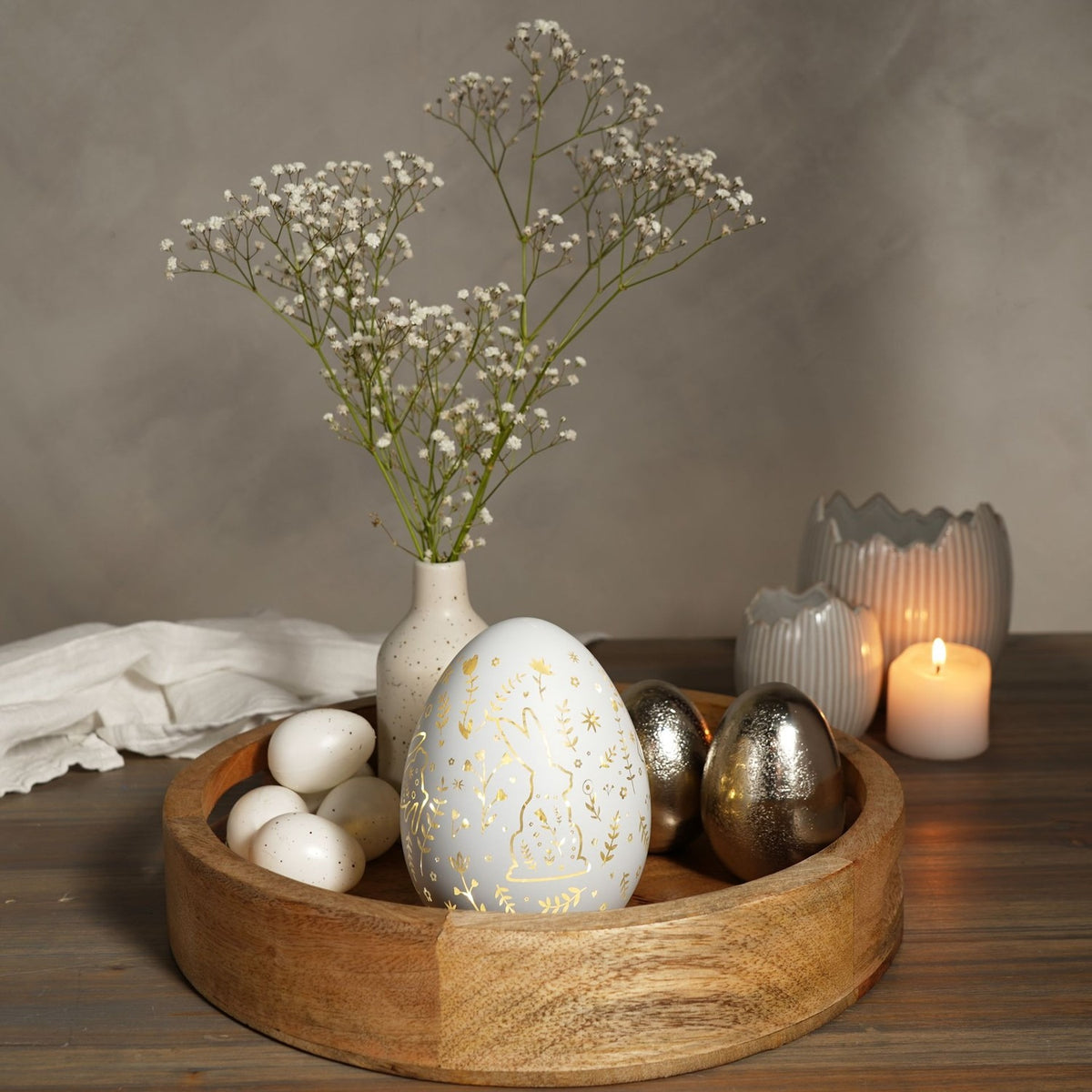 White glass Easter egg light-up decoration with gold rabbit and floral pattern, styled in a wooden bowl with eggs, gypsophila in a vase, and candles.