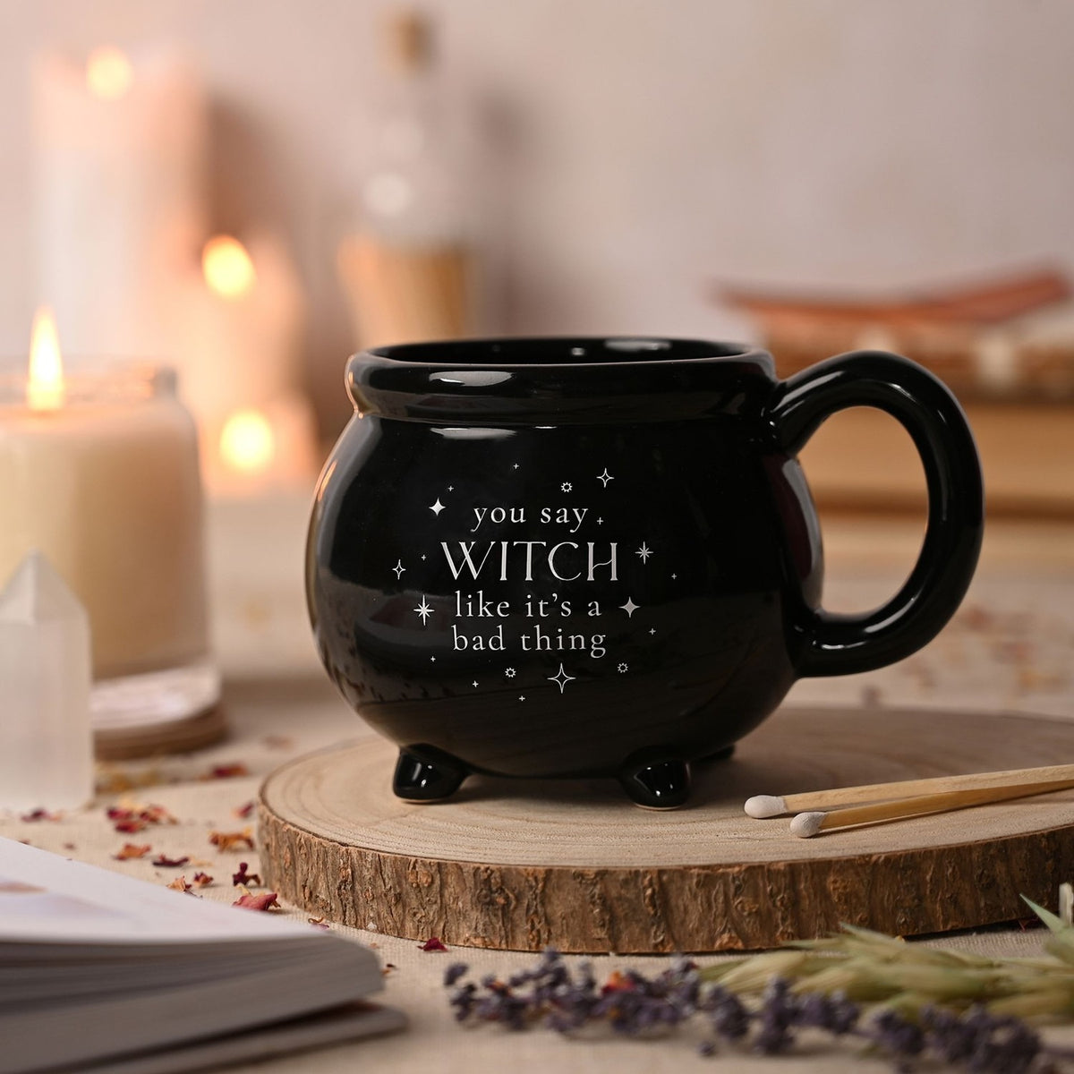 Black ceramic cauldron mug with stubby feet and loop handle, reading "you say WITCH like it's a bad thing" in white lettering, shown beside candles and a crystal.