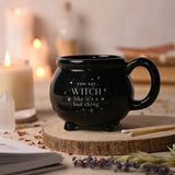 Black ceramic cauldron mug with stubby feet and loop handle, reading "you say WITCH like it's a bad thing" in white lettering, shown beside candles and a crystal.