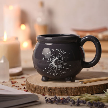 Purple ceramic cauldron-shaped mug with rolled rim, stubby feet, and white "Pick Your Poison" moon and stars print, shown on a wooden coaster in a candlelit setting.