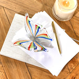 Clear glass butterfly figurine with open wings in rainbow stripes of red, orange, green and blue, displayed on white paper beside a gold pen and candle on a wooden desk.