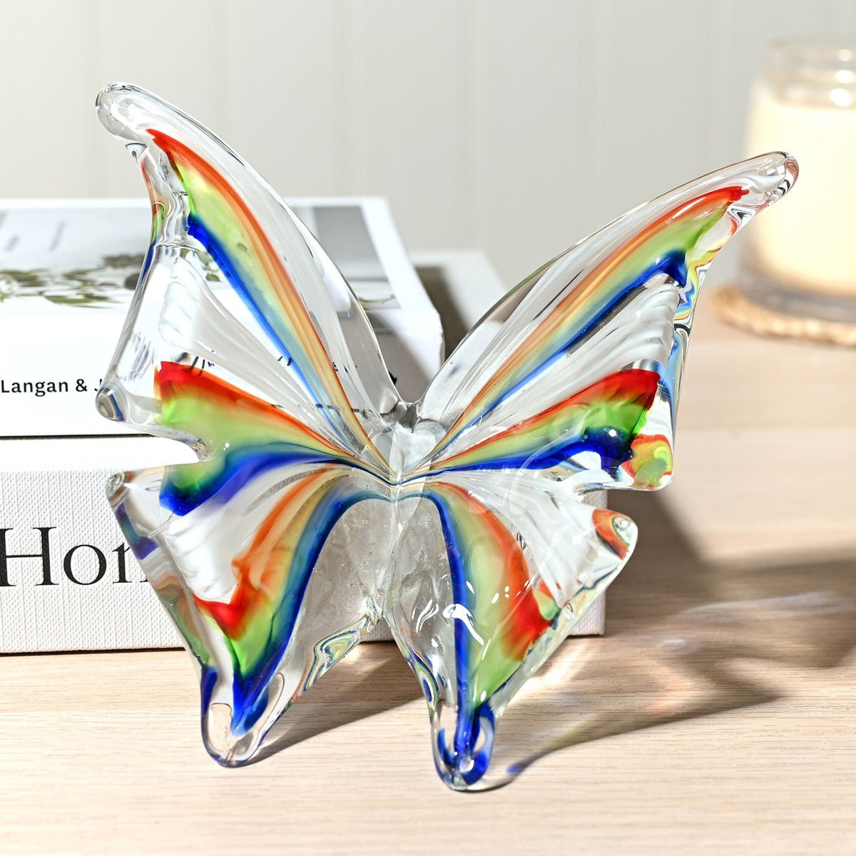 Clear glass butterfly figurine with open V-shaped wings striped in red, orange, green and blue, displayed on a wooden surface beside books and a candle.