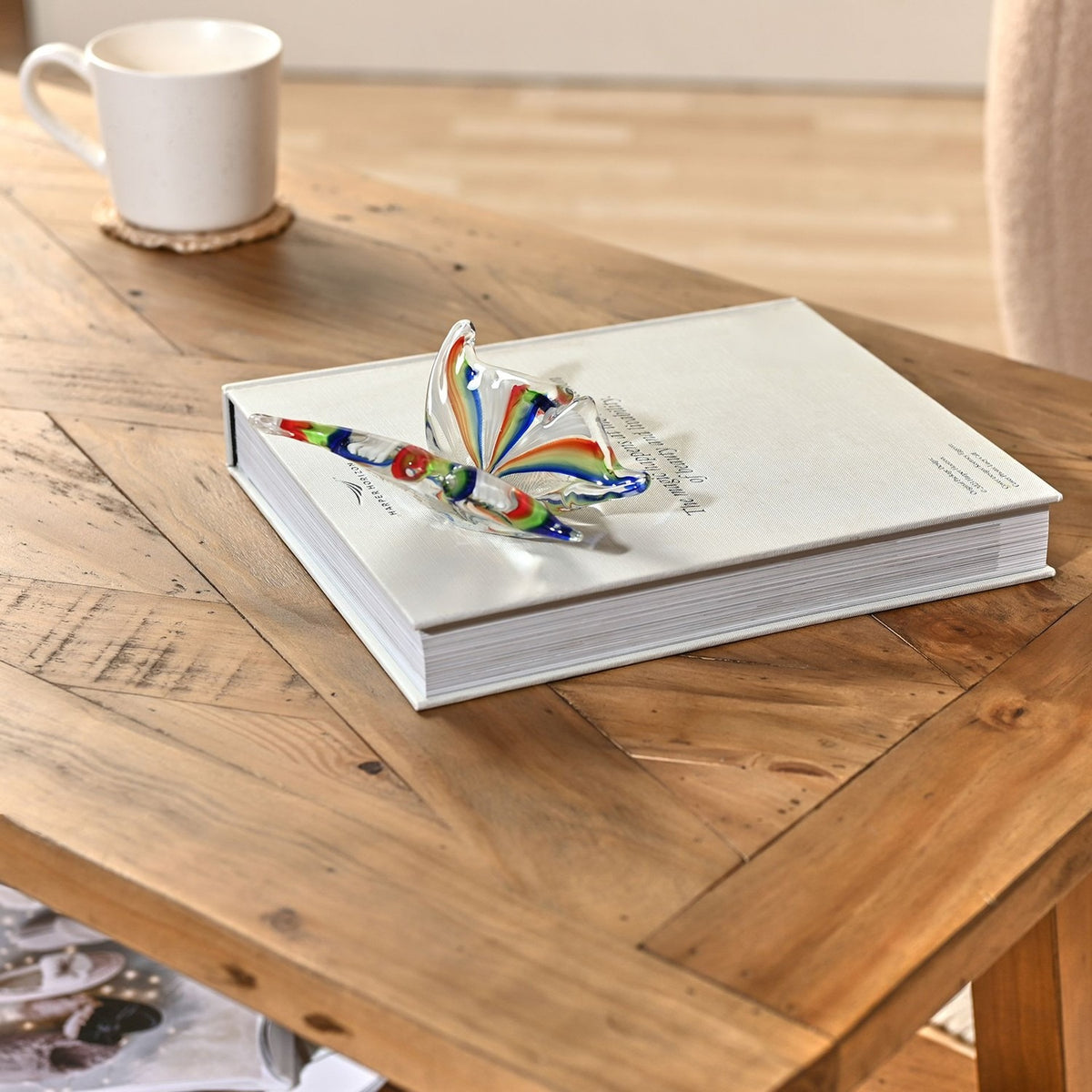 Rainbow Striped Open Wing Butterfly Glass Figurine in clear glass with red, orange, green and blue stripes, displayed on a white book atop a wooden coffee table.