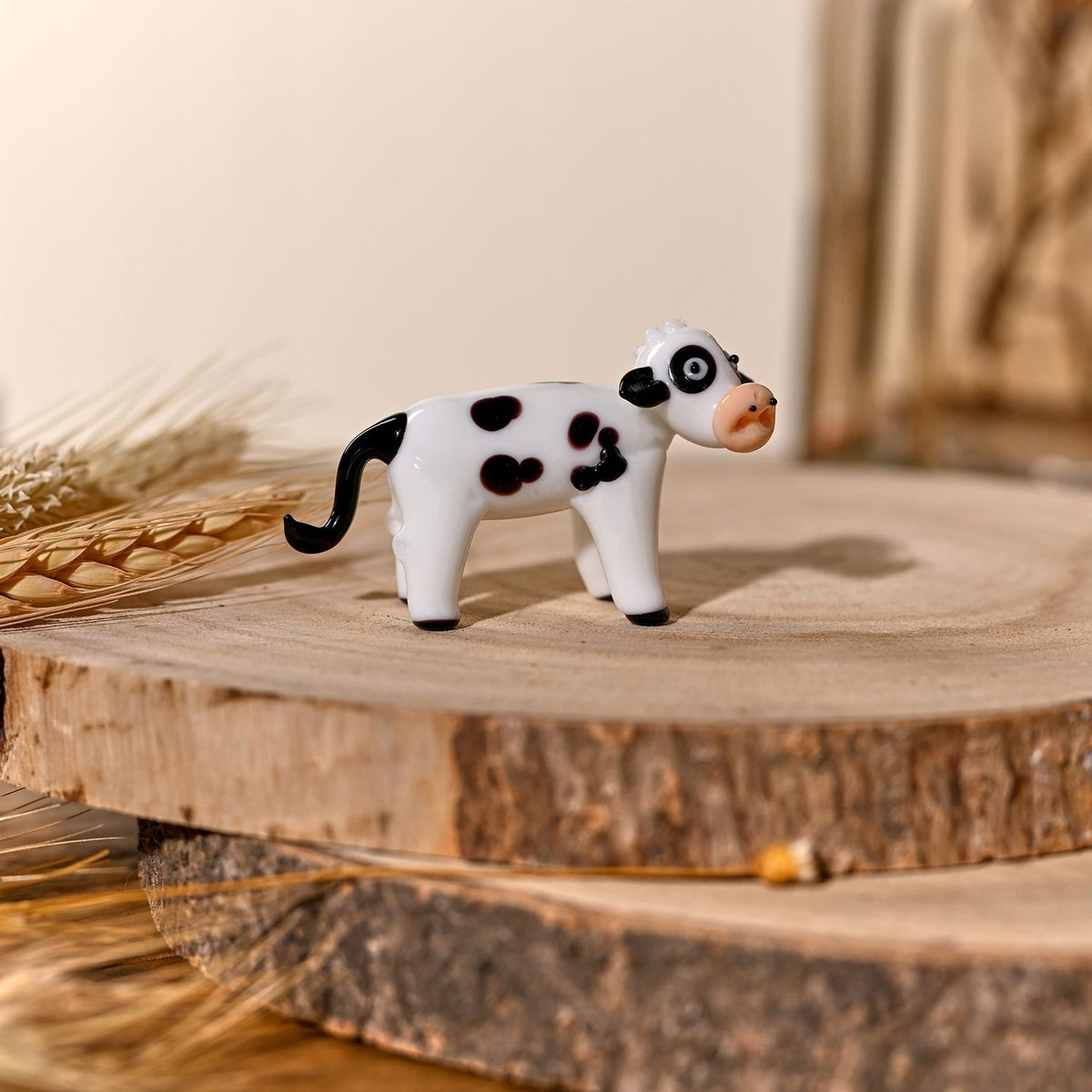 Standing Black Spotted Cow Glass Miniature Figurine in milky white glass with black spots, peach muzzle and curved tail, displayed on a wood slice with dried wheat stalks.