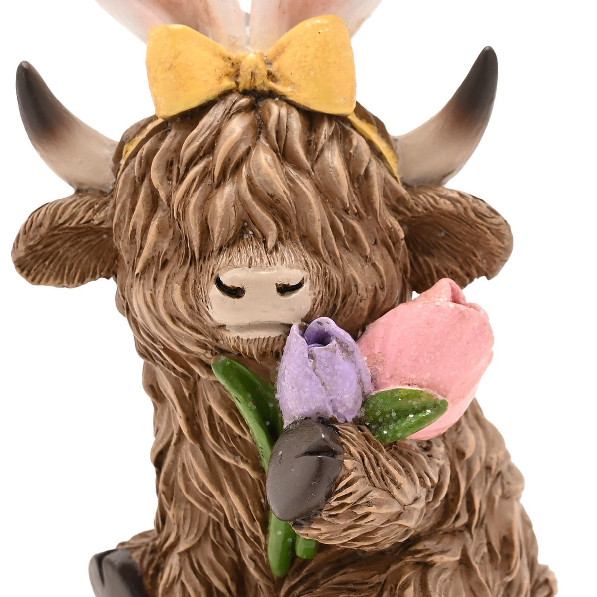 Close-up of a resin Highland cow Easter decoration in brown, wearing white bunny ears tied with a yellow bow and holding a lilac and pink tulip posy near its nose.