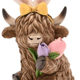 Close-up of a resin Highland cow Easter decoration in brown, wearing white bunny ears tied with a yellow bow and holding a lilac and pink tulip posy near its nose.
