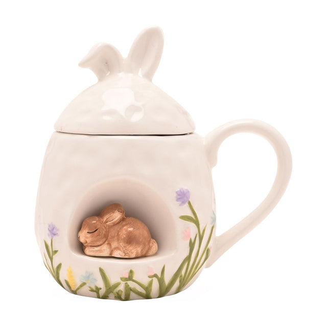 White ceramic Easter mug with bunny-ear lid and a 3D sleeping bunny in an arched nook, painted grass and pastel flowers, shown on a plain white background.