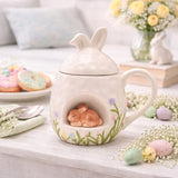 White ceramic Easter mug with bunny-ear lid and 3D sleeping bunny in an arched nook, painted grass and pastel flowers, shown on a table with Easter decor.
