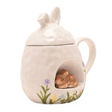 White ceramic mug with bunny-ear lid and a 3D sleeping bunny nestled in a front nook, painted with grass and pastel floral details, shown on a plain background.
