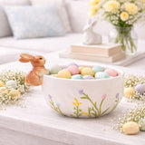 White round ceramic bowl with a 3D sandy brown bunny on the rim and pastel wild flowers, shown filled with speckled eggs on a styled Easter table.