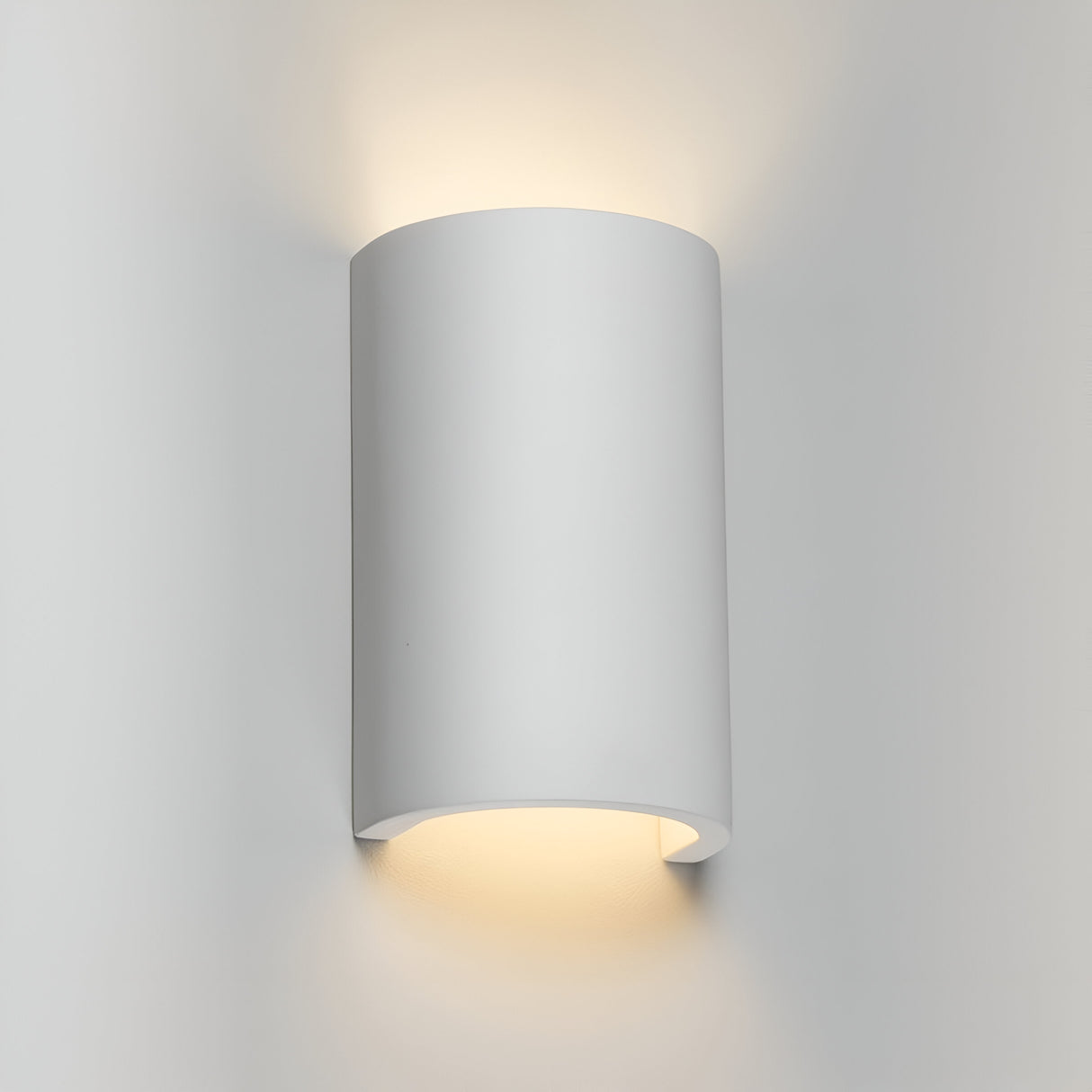 The Curved Up And Down Plaster Wall Light casts gentle illumination both upwards and downwards on a neutral wall, establishing a cozy and contemporary ambiance.