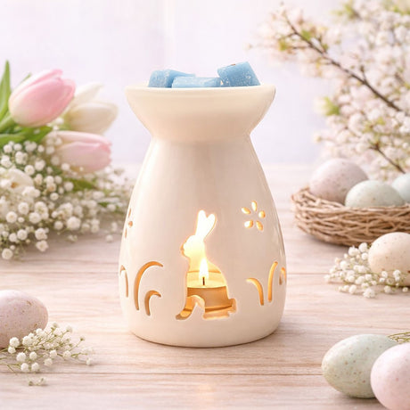 Cream ceramic wax melt burner with a bunny silhouette, flowers and grass cut-outs glowing from a lit tealight, blue wax melts on top, shown among Easter décor.
