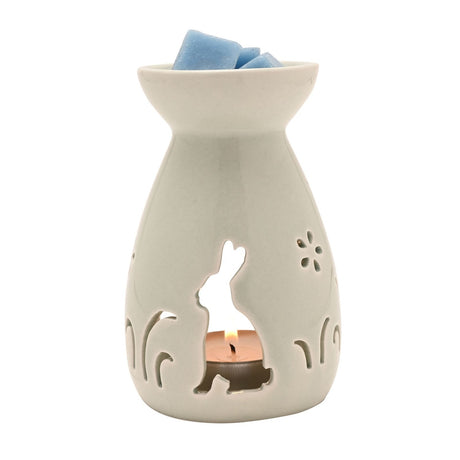 Ceramic wax melt burner in white with a cut-out sitting bunny, flower and grass silhouettes, lit tealight glowing inside, topped with blue wax melts, on a plain white background.