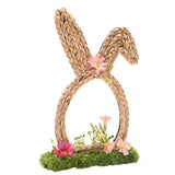 Faux flower bunny ears decoration in natural woven braid with egg-shaped loop, pink faux daisy accents, and a mossy green base, shown on a white background.