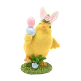 Yellow resin Easter chick figurine with bunny ears and floral band, holding pink and blue painted eggs, standing on a grassy base against a plain white background.