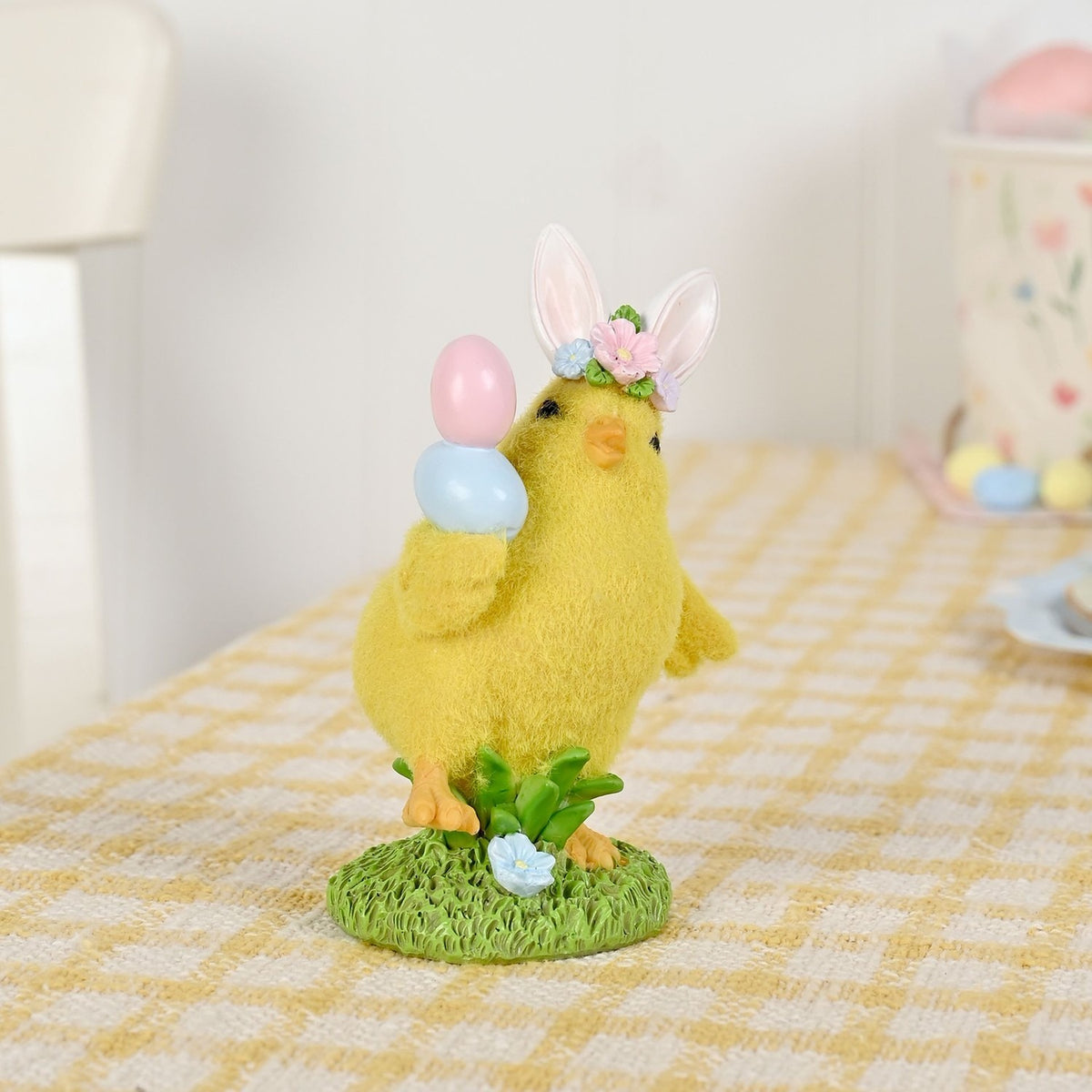 Bunny Ear Easter Chick Resin Figurine in yellow with fuzzy texture, pink floral ear band, orange beak and feet, holding pink and blue eggs on a grassy base.