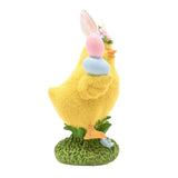 Yellow resin Easter chick figurine with bunny ears and floral band, holding pink and blue eggs, fuzzy textured body on a green grass base, plain white background.