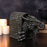 Resin figurine of a roaring grey dragon with clawed foot breaking through a brick archway, shown with lit candles and a dark cauldron in a dimly lit setting.