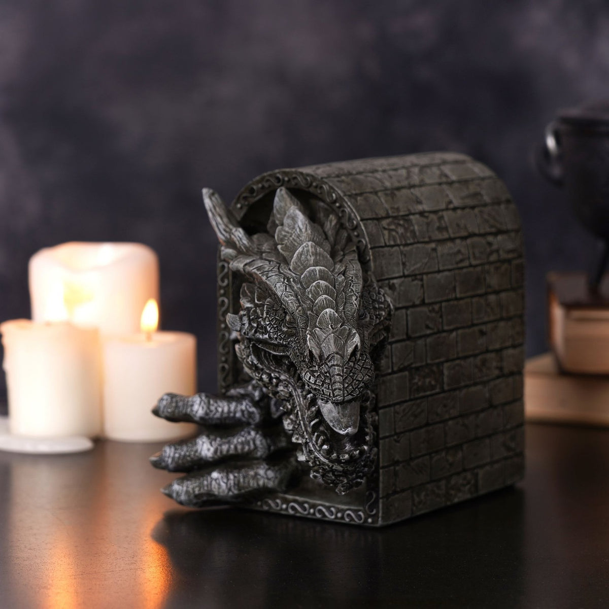 Grey resin figurine of a roaring dragon with clawed foot breaking through a brick archway, set on a dark surface beside lit candles and stacked books.