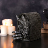 Grey resin figurine of a roaring dragon with clawed foot breaking through a brick archway, set on a dark surface beside lit candles and stacked books.