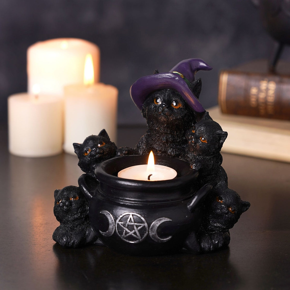 Black resin tealight candle holder featuring five amber-eyed cats around a cauldron with a pentagram emblem, the largest wearing a purple witch's hat, shown lit beside white candles.
