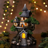 LED Treehouse Resin Light-Up Decoration, 19.8cm, grey slate-style turreted roofs and stone doorway with green foliage, illuminated windows, on a dark surface with blurred fairy lights.