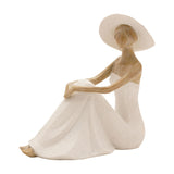 Floor seated lady resin figurine in cream, wearing a wide-brimmed hat and textured strapless gown, knee drawn up with arms folded, natural wood-tone skin and hair, on a plain white background.