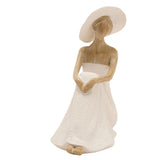 Floor seated lady resin figurine with wide-brimmed hat, seated pose with knee drawn up and arms folded, cream textured gown, natural finish on face and hair, on a plain white background.