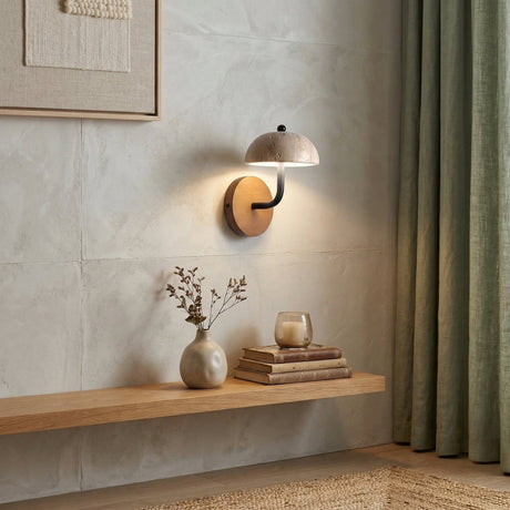 The Marvella LED Dome Travertine Wall Light in walnut brown and light beige brightens a minimalist wooden shelf with a ceramic vase, glass candle, and stacked books; green curtains hang nearby.