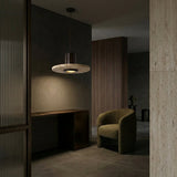 A modern, minimalist room with dark walls and flooring features a curved green armchair, sleek brown desk, ribbed wooden panels, and is illuminated by the Torvane 1 Light Large Wide Disc Travertine Single Pendant Light in walnut brown and light beige.