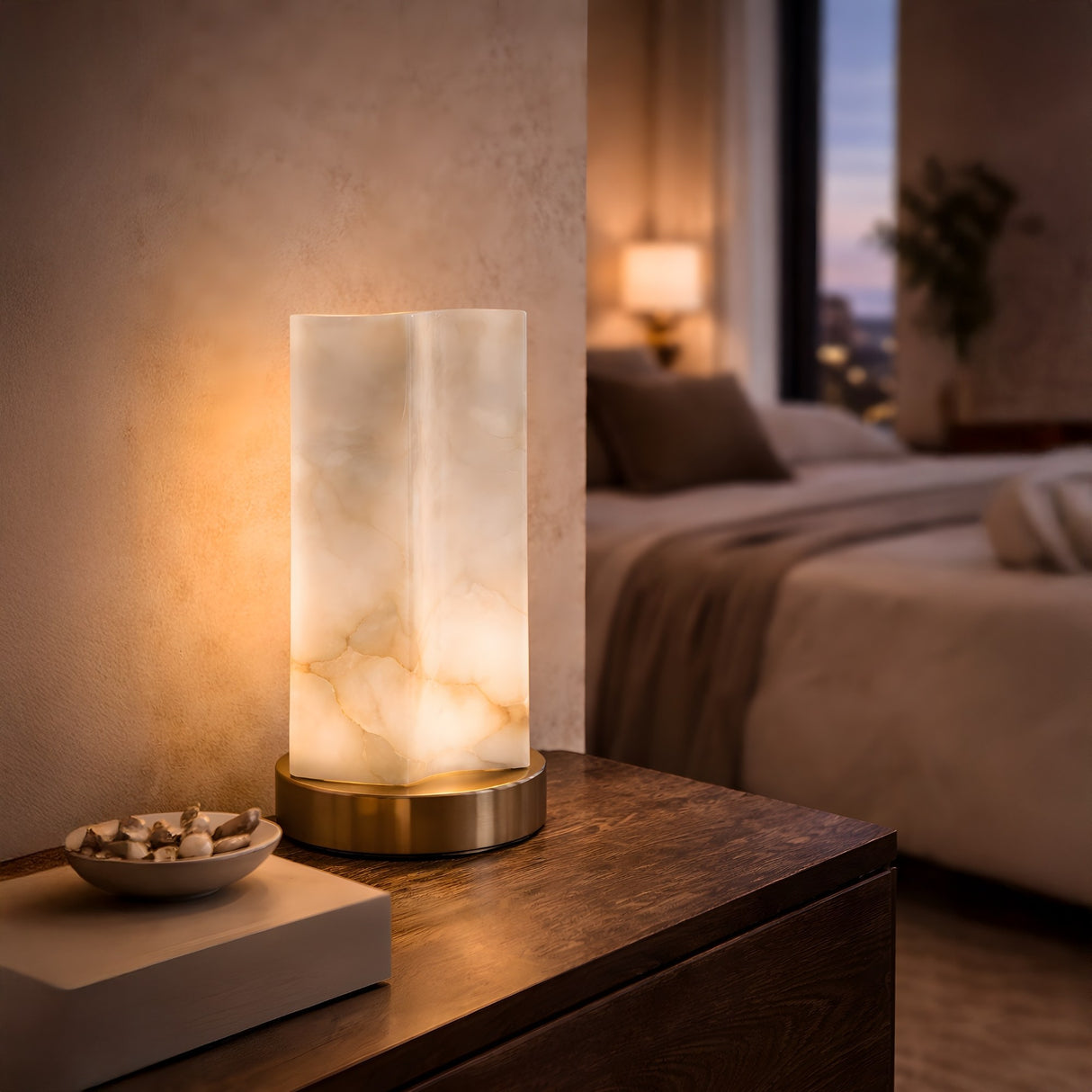 The Virelle LED Curved Panel Alabaster Table Lamp - Soft White sits on a wooden nightstand beside a decorative bowl in a softly lit, modern bedroom with a neatly made bed and warm ambient lighting.