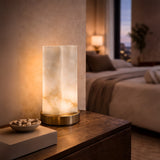 The Virelle LED Curved Panel Alabaster Table Lamp - Soft White sits on a wooden nightstand beside a decorative bowl in a softly lit, modern bedroom with a neatly made bed and warm ambient lighting.