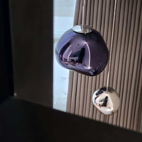A close-up of two Lava Medium LED Pendant Lights—one in Smoked Blue and one in silver—hanging before beige vertical blinds, ideal for contemporary interiors with a window framed in dark tones partially visible.
