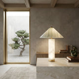 A modern, minimalist room with a marble table featuring the Veridra 1 Light Conical Shade Alabaster Table Lamp in Soft White & Green, stacked books, and a sculpture. A large window reveals a bonsai tree in a tranquil courtyard of neutral tones.
