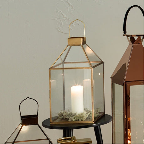 Shiny Brass & Glass Lantern, 49cm tall pyramid-shaped candle holder with peaked handle, glass panels and a white pillar candle, shown on a black side table.