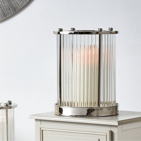 Silver stainless steel and clear glass rod hurricane lamp, 33.5cm, with a lit candle inside, shown on a white cabinet against a light grey wall.