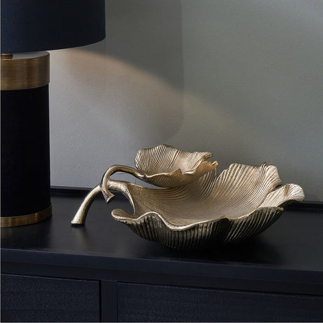 Gold Aluminium Dual Gingko Leaf Bowl, 37cm, with textured overlapping leaf-shaped tiers and a curved stem base, shown on a dark sideboard beside a black table lamp.