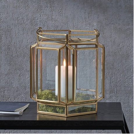 Shiny Brass & Glass Hexagon Wide Lantern, 28cm, with glass panels and a white pillar candle and moss inside, shown on a dark table against a grey textured wall.