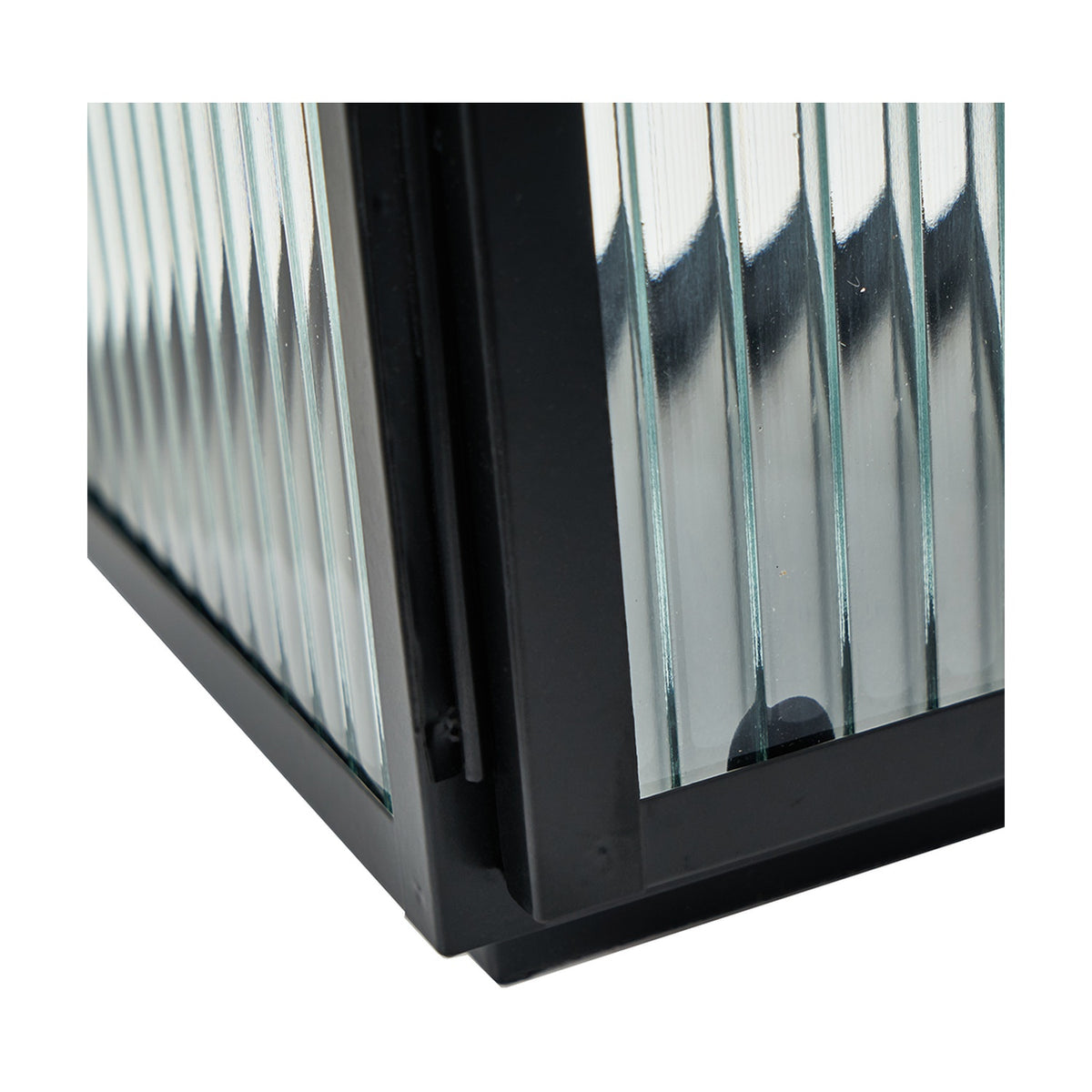 Close-up corner detail of a black iron candle lantern with vertical ribbed glass panels, showing the metal frame joints against a plain white background.