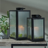Set of 2 black iron and ribbed glass lanterns with curved top handles, each holding a lit candle, shown on a wooden table beside a potted plant.