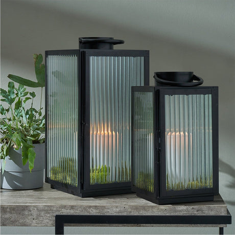 Set of 2 black iron and ribbed glass lanterns with curved top handles, each holding a lit candle, shown on a wooden table beside a potted plant.