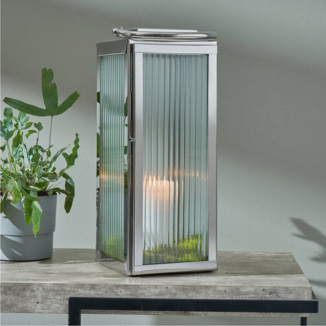 Silver stainless steel and ribbed glass small lantern, 43cm, with a lit candle inside, shown on a wooden table beside a potted plant against a light grey wall.