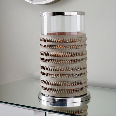 Large hurricane lamp with clear glass cylinder, silver metal rims, and grey plaited leather banding, displayed on a mirrored table against a light grey wall.