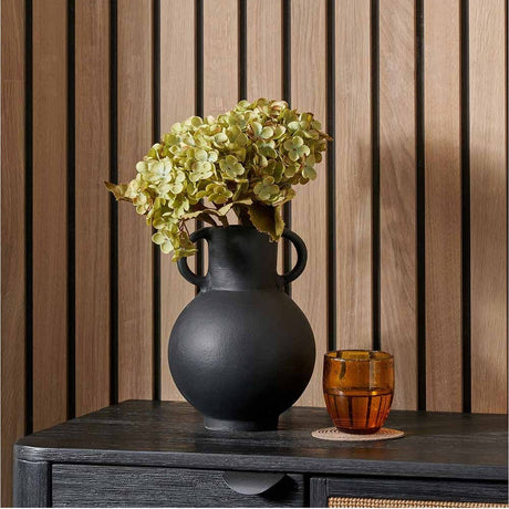 Matt Black Aluminium Vase with Handles - 24cm holding green hydrangea stems on a dark sideboard, with an amber glass tumbler beside it against a wood-slat wall.